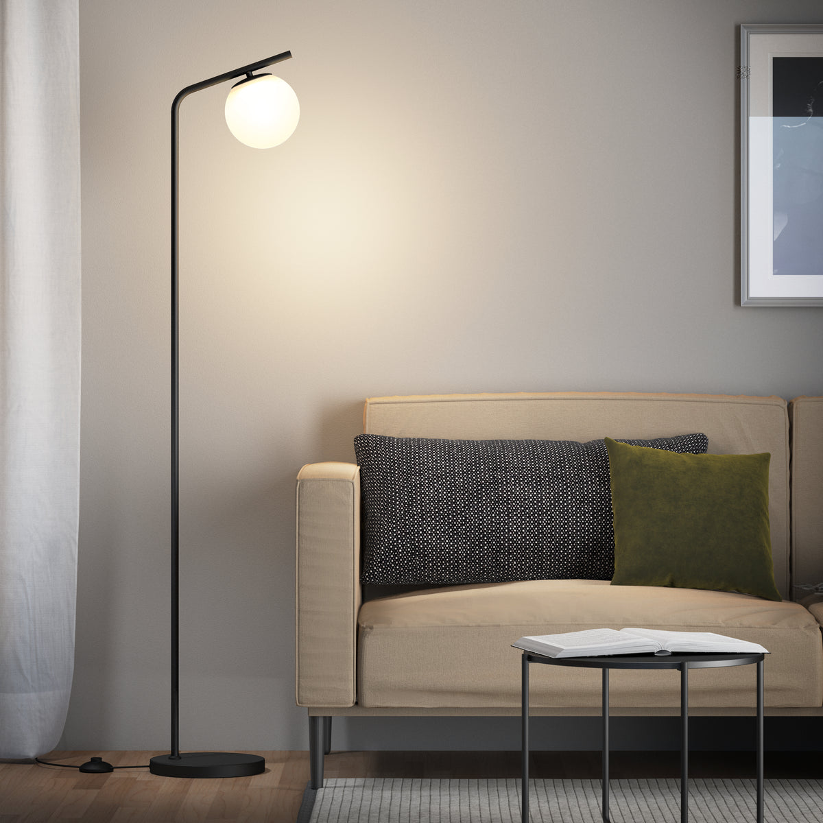 A modern living room with a beige sofa, black and green cushions, a round floor lamp with a glowing white globe, a small table with an open book, and a framed picture on a light-colored wall.