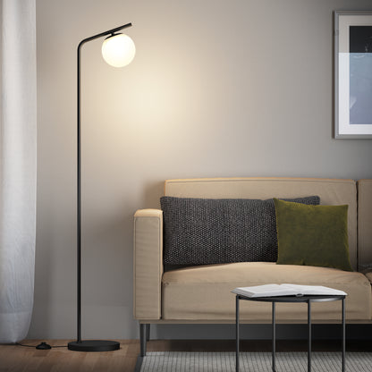 A modern living room with a beige sofa, dark and green throw pillows, a round black side table holding an open book, and the Artika Clayton dimmable LED modern mid-century floor lamp with a glowing round white shade beside the sofa.