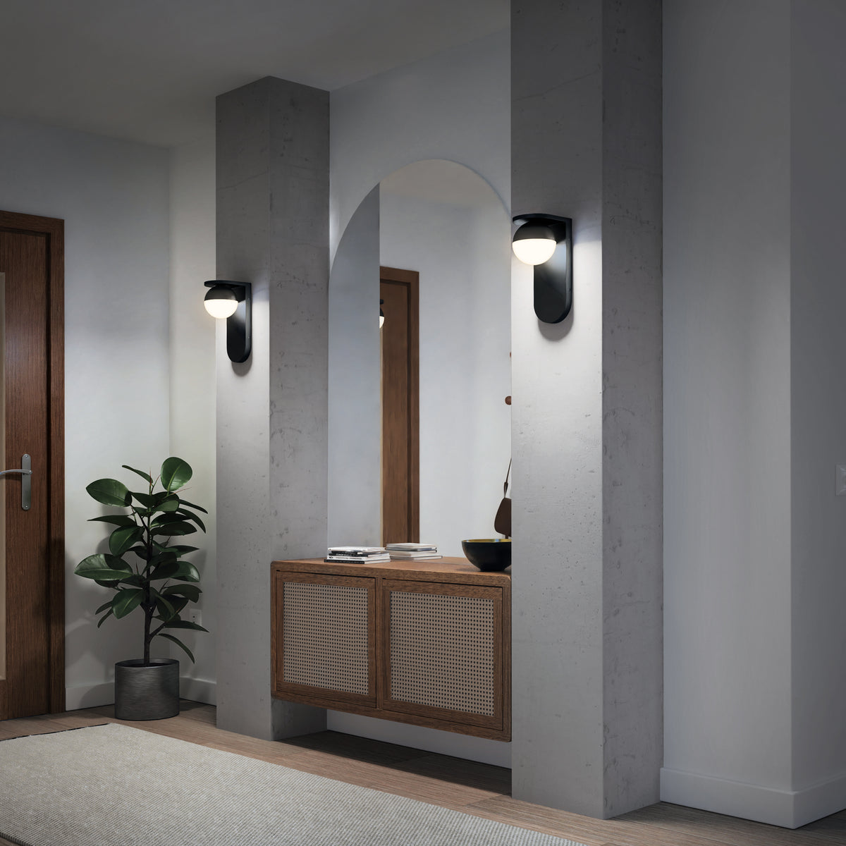 A modern entryway featuring Artika’s Bloom indoor-outdoor LED wall light, a large arched mirror, concrete columns, a wooden console with woven paneling, a potted plant, and a closed wooden door in the background.