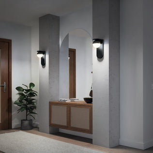 A modern entryway featuring Artika’s Bloom indoor-outdoor LED wall light, a large arched mirror, concrete columns, a wooden console with woven paneling, a potted plant, and a closed wooden door in the background.