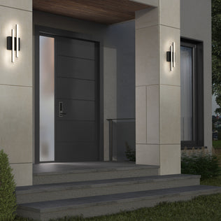 Modern house entrance with wide dark gray door, frosted side window, concrete steps, beige walls, and two Artika Grid indoor-outdoor LED wall lights on either side of the door. Greenery appears in the lower left corner.