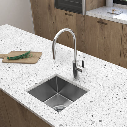 A modern kitchen features the Artika Plaza Bar Single Bowl Sink with a curved faucet and a wooden cutting board holding chopped green onions on a white speckled countertop, complemented by wood cabinets in the background.