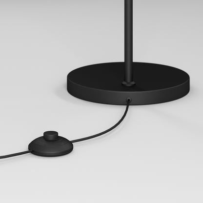 A close-up of the Artika Clayton dimmable LED modern mid-century floor lamp’s black base with a matching cord and round foot switch, shown on a white surface.