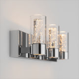 A modern wall sconce with three cylindrical glass shades, featuring tiny bubble-like details and warm LED lights, mounted on a sleek, reflective chrome base against a light-colored wall.