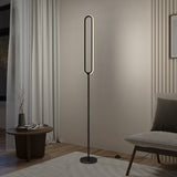 A modern, oval-shaped LED floor lamp with a black stand is lit in a minimalist living room, next to a small round table, a light gray chair, and beige curtains.