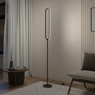 The Artika Leni dimmable LED modern floor lamp, featuring an oval design and black stand, emits soft light in a minimalist living room beside a small round table, light curtains, and a cushioned chair.