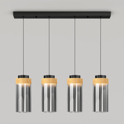 The Artika Hanson 4-light LED pendant features cylindrical glass shades, black fixtures, and light wood accents, suspended in a row from a rectangular black ceiling mount against a plain light gray background.