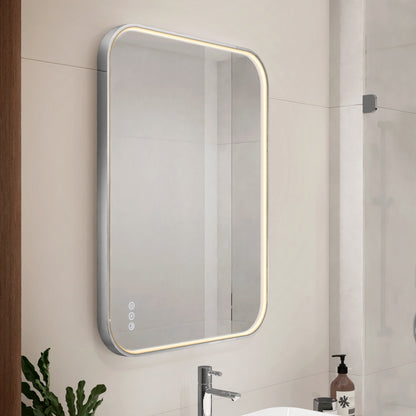 A rectangular bathroom mirror with rounded corners and built-in LED lighting along the edges is mounted above a sink. Touch controls are visible on the mirror, and a soap dispenser and plant are nearby.