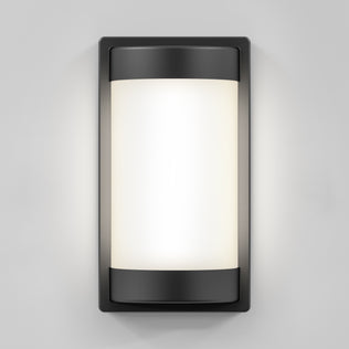 The Artika Fogo modern LED indoor-outdoor wall light 5 CCT features a frosted cylindrical glass panel and black metal frame, casting a soft white glow against a light gray wall.