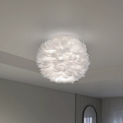 The Artika Plume LED flush mount light features layered, feather-like materials and emits a soft glow, adding modern elegance to a minimalist room with light walls and a round mirror.