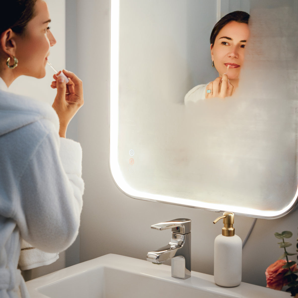 A woman in a white robe applies lipstick before the Artika Royce 24″x32″ dimmable anti-fog LED mirror.