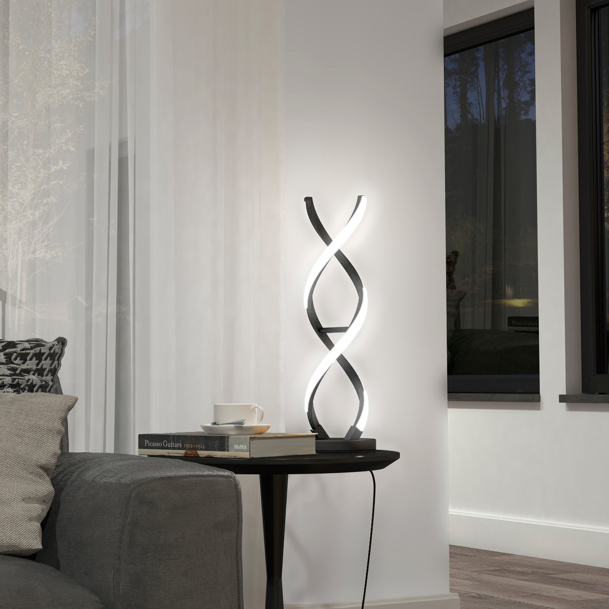 A modern spiral LED table lamp glows on a black round side table beside a gray sofa. The table holds books and a white cup. Large windows and sheer curtains are in the background.