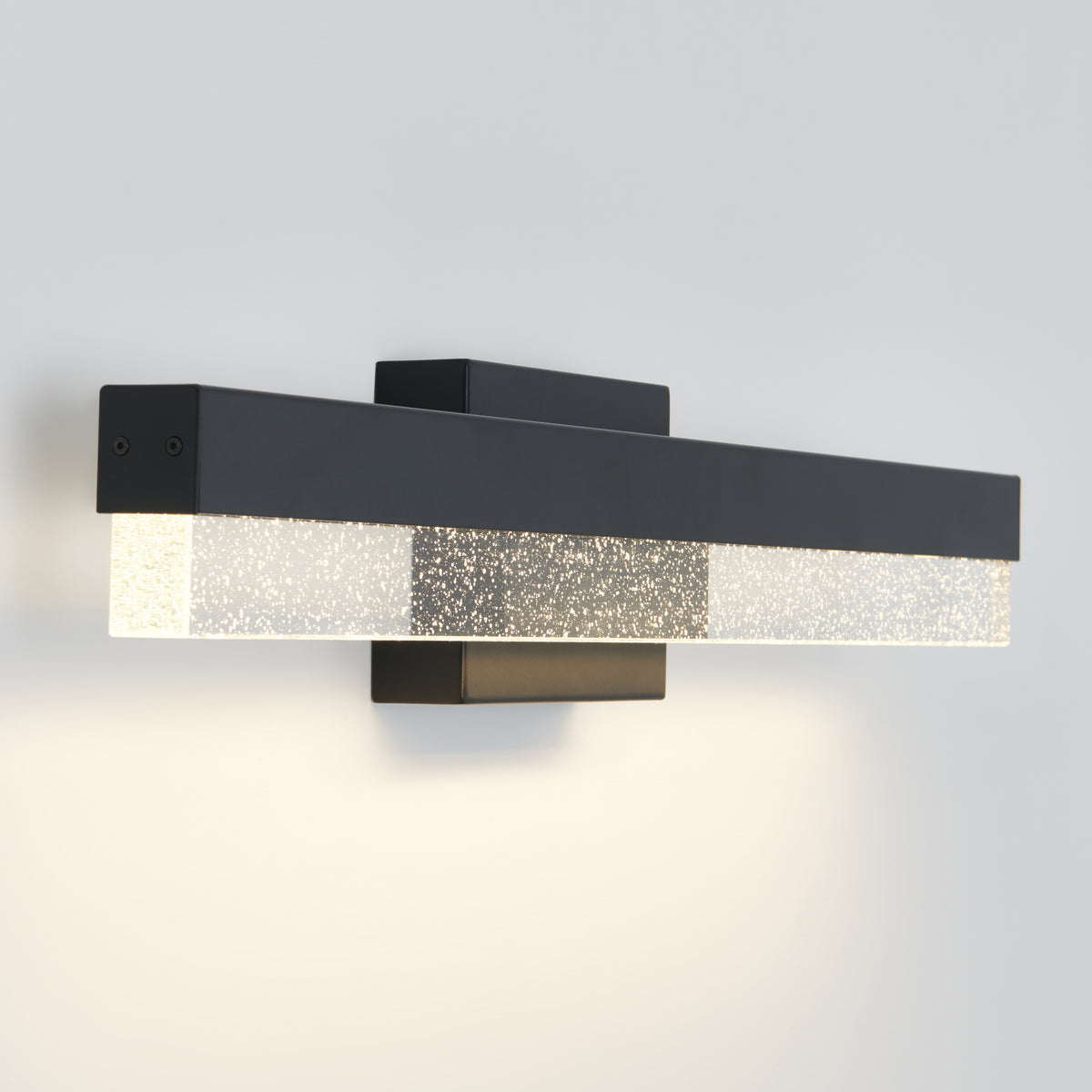 A modern black wall sconce with a rectangular LED light, featuring a frosted, textured glass cover that emits a soft, even glow against a plain white wall.