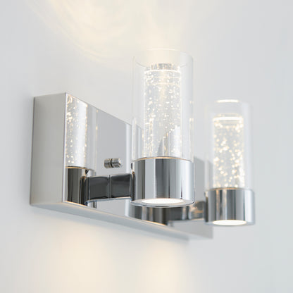 The Artika Ratio 2-light LED bathroom vanity light features two bubble-filled cylindrical glass shades on a chrome rectangular base, beautifully illuminated against a white wall.
