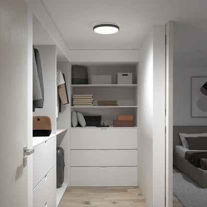 A modern walk-in closet with white shelves, drawers, and organized clothes is illuminated by the Artika Sunraker 10 in. LED modern flush mount light; a softly lit bedroom with a bed and wall art is visible through the open door.