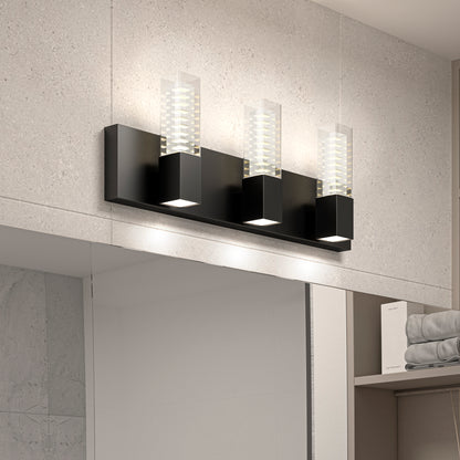 The Artika Hologram LED modern bathroom vanity light 5 CCT, featuring three rectangular black lights with clear glass shades, is mounted above a bathroom mirror on a light gray tiled wall with shelves of neatly folded towels nearby.