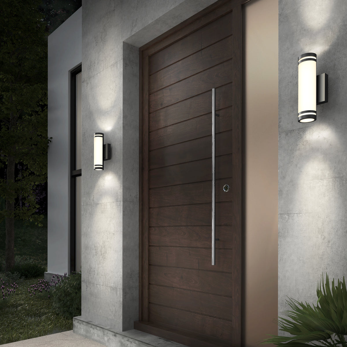 Modern wooden front door with horizontal panels, a tall metal handle, and two cylindrical wall lights on either side, mounted on a concrete exterior wall. Green foliage is visible in the foreground and background.