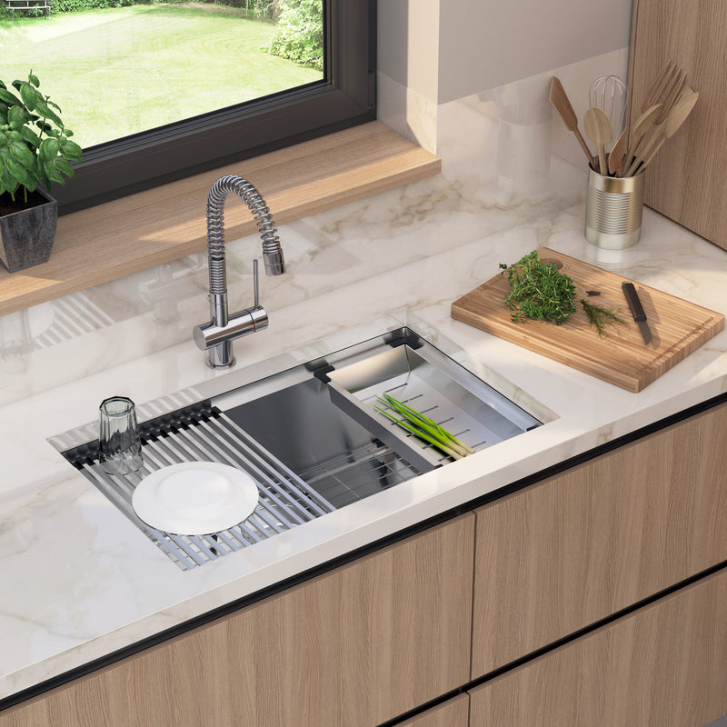 A modern kitchen features the Artika Newport 31 in. stainless steel workstation sink with a flexible faucet and built-in drying rack for dishes and green onions. A cutting board with herbs and utensils sits nearby by the window.
