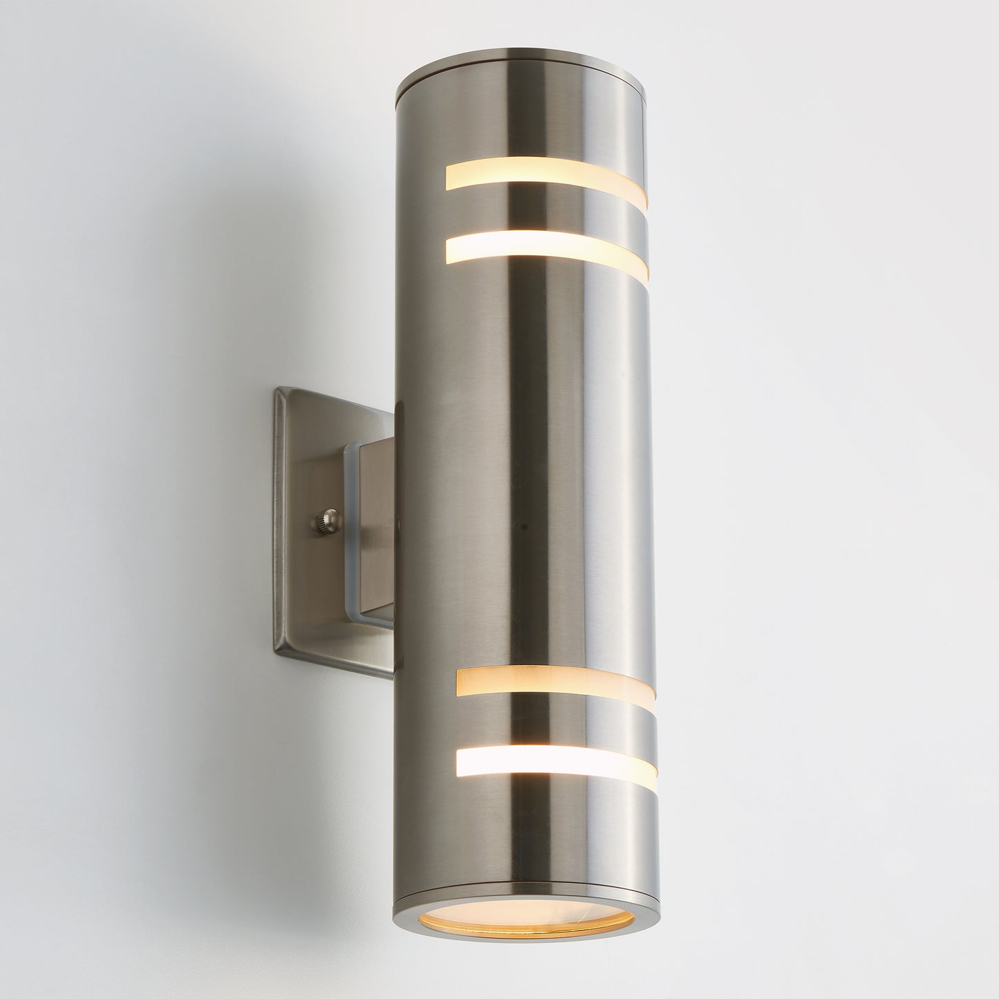 The Artika Voltz indoor-outdoor wall light is a modern cylindrical sconce with a brushed metal finish, mounted on a square base. It has horizontal cutouts shining warm light from top and bottom, shown against a plain white background.