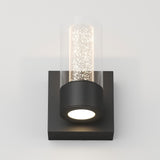 A modern wall sconce with a cylindrical glass shade featuring small bubbles, mounted on a black square base, emitting warm white light.