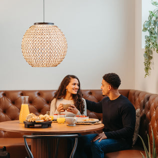 A smiling couple enjoys breakfast at a round wooden table in a cozy dining nook, illuminated by the Artika Benson LED pendant light 5 CCT hanging overhead.