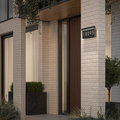Modern house entrance with light-colored brick walls, a wooden door, large windows with white curtains, a black planter with green shrubs, and an illuminated address sign displaying the number 18095.