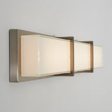 A modern, rectangular wall sconce with a brushed metal base and three frosted glass panels emitting soft, warm light, mounted on a plain white wall.
