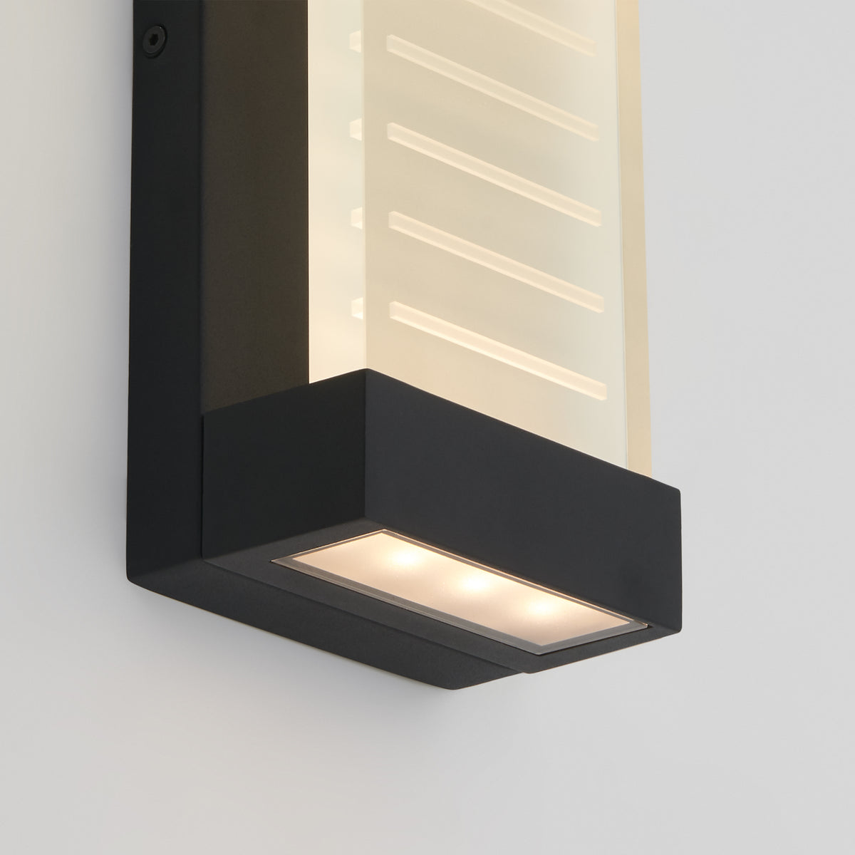 A modern wall sconce with a black rectangular frame and frosted panel diffuser, mounted on a white wall, emitting soft light from the bottom.