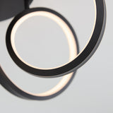 Close-up of a modern ceiling light fixture featuring two intertwined black metal rings with integrated LED lights, glowing softly against a plain light gray background.