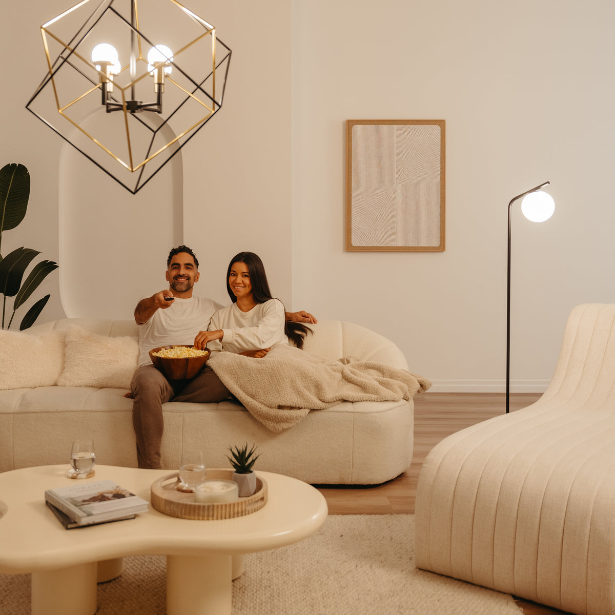A couple sits on a cozy cream-colored sofa, smiling and watching TV while sharing a bowl of popcorn in a modern, warmly lit living room with stylish decor and minimalist furniture.