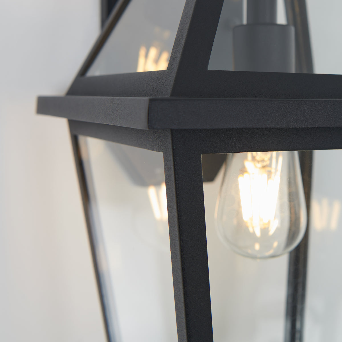 Close-up of a modern black metal and glass outdoor wall lantern with a visible glowing filament light bulb inside.