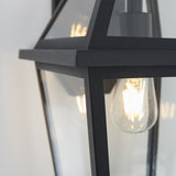 Close-up of a modern black metal and glass outdoor wall lantern with a visible glowing filament light bulb inside.