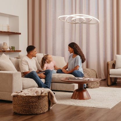 A family of three sits on a beige couch in a modern living room with warm lighting, neutral tones, and contemporary decor, highlighted by the Artika Swirl Butterfly LED contemporary pendant light 3 CCT overhead.
