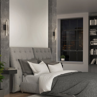 Modern bedroom with a tufted gray headboard, gray and white bedding, Artika Grid indoor-outdoor LED wall lights on either side, shelves holding books and decor, and a large window overlooking a nighttime city skyline.
