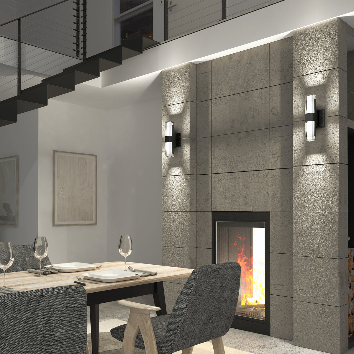 A modern dining area with a wooden table set for four, gray upholstered chairs, a concrete fireplace with a burning fire, wall sconces, and a staircase with a metal railing above.