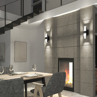 Modern dining room with a wooden table for four, gray upholstered chairs, and a concrete fireplace. Artika’s Gemini indoor-outdoor LED wall lights illuminate the space beneath an open staircase adorned with artwork.