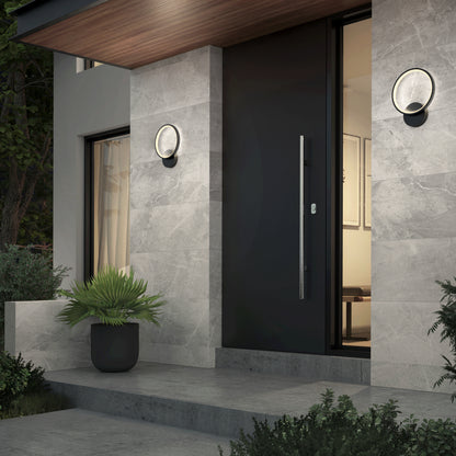 Modern house entrance featuring a large black front door, gray stone walls, a potted plant, and two Artika Solis modern LED indoor-outdoor wall lights (5 CCT). The door is partially open with shrubs and steps leading to the softly lit interior.