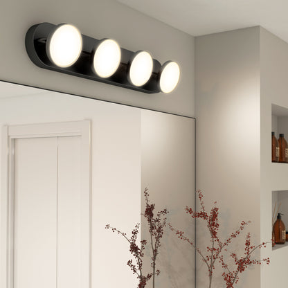 A modern bathroom features the Artika-CAN-dev Santori LED bathroom vanity light with a matte black finish and four round bulbs above a mirror, white walls, and shelves displaying bottles and vases.