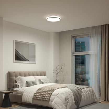 A modern bedroom features a beige upholstered bed, white and gray bedding, a side table with a branch in a vase, wall photo, city-view window with sheer curtains, and Artika’s Hemisphere 15 in. LED flush mount light with adjustable brightness.