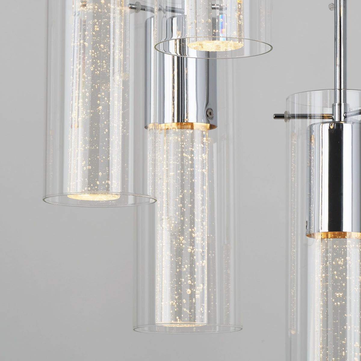 Close-up of a modern chandelier featuring cylindrical glass shades with tiny bubbles and chrome accents, each cylinder illuminated with soft, warm light. The background is a plain light gray.
