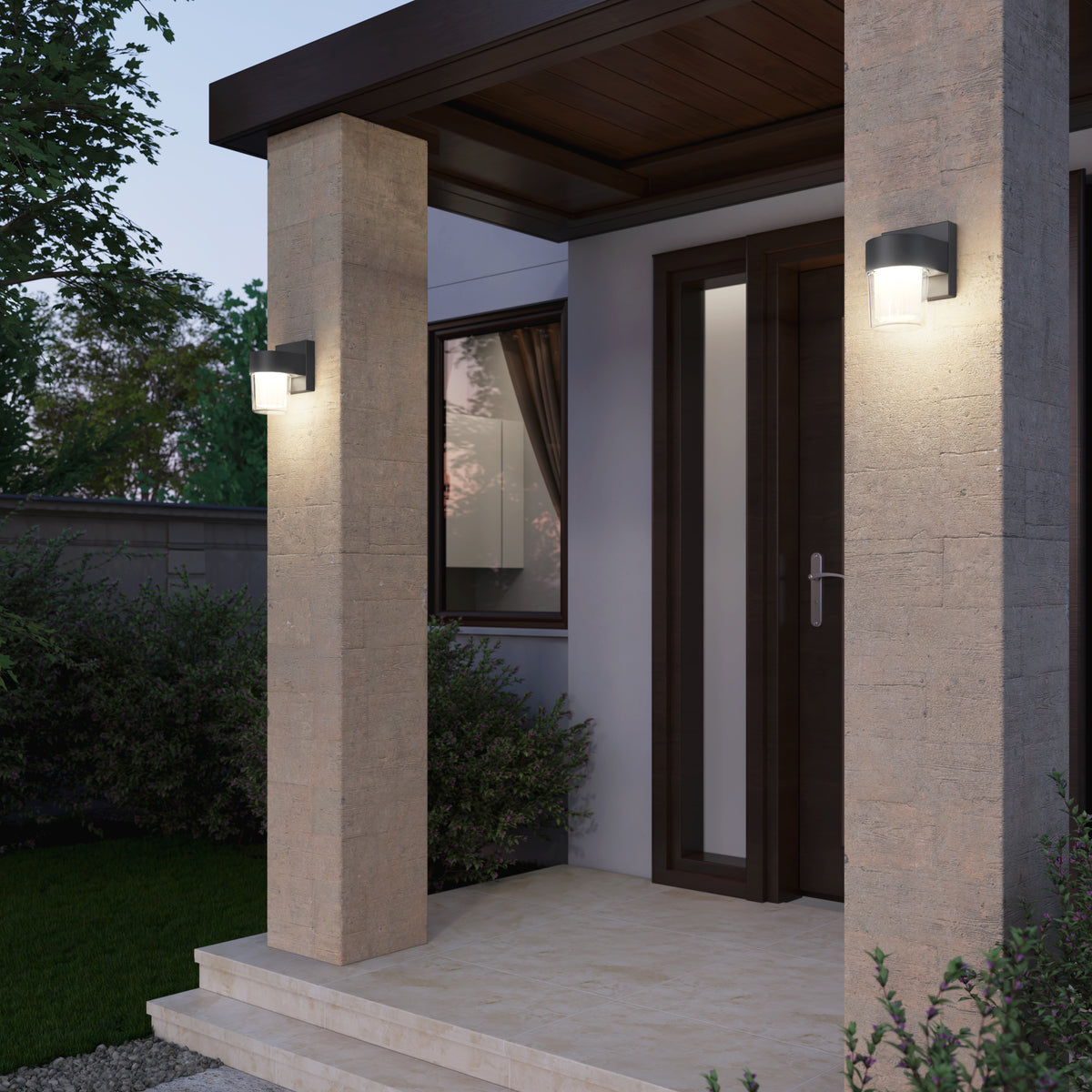 Modern front porch with two wall-mounted outdoor lights on stone pillars, a wooden door with glass panels, and surrounding greenery at dusk.
