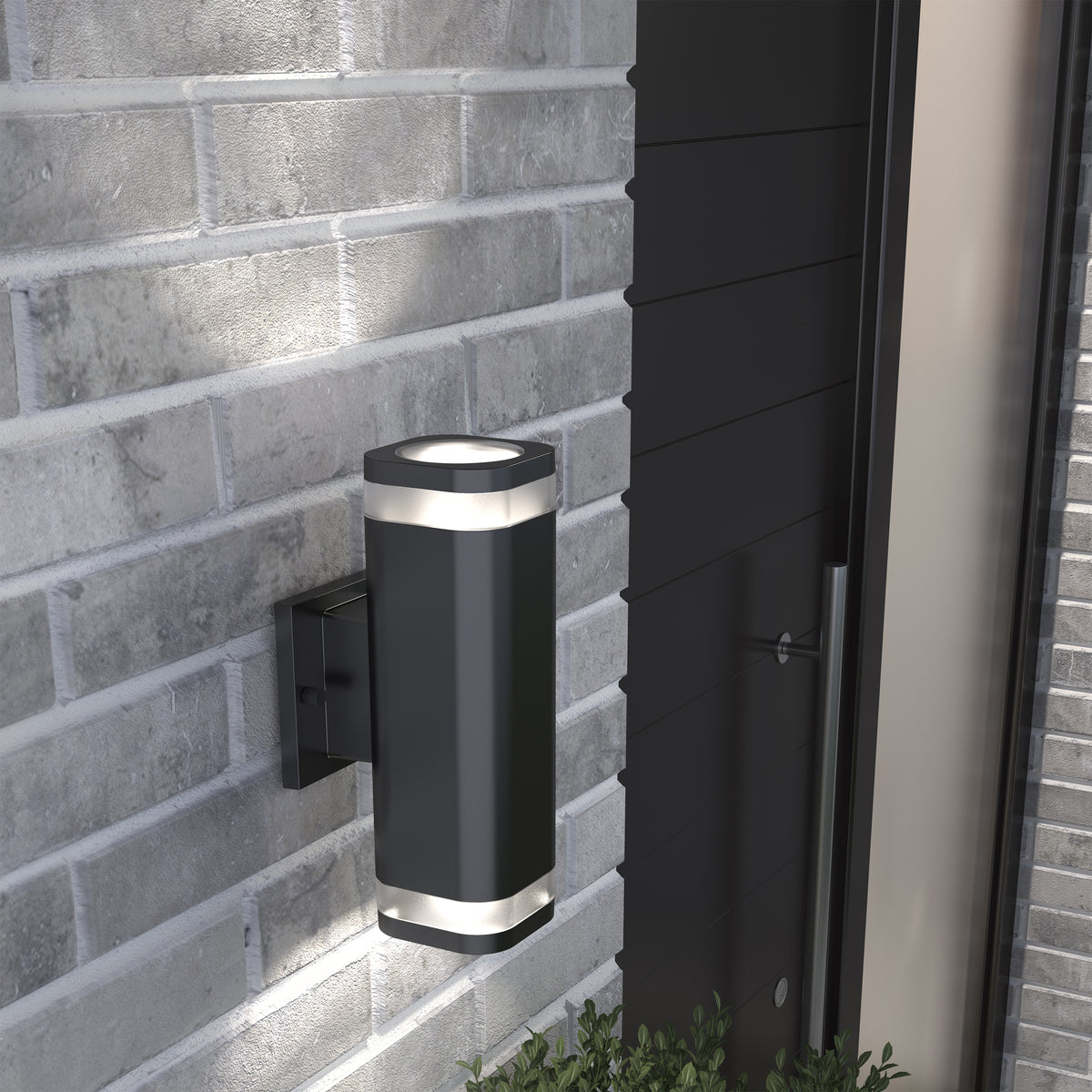 A modern black outdoor wall light fixture with two illuminated ends is mounted on a light gray brick wall next to a dark door. Green plants are visible below the fixture.