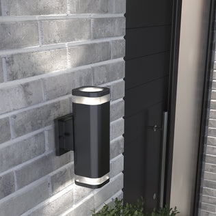 The Artika Mettle LED indoor-outdoor wall light, featuring a modern rectangular black design, is mounted on a gray brick wall near a dark door with a plant below.