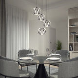 A modern dining room with a round glass table set for four, gray upholstered chairs, contemporary pendant lights above, sheer curtains, and a bookshelf with decor and a potted plant in the background.