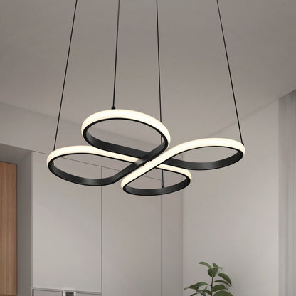 A modern, black and white LED chandelier with looping, intertwined rings hangs from the ceiling in a minimalist room with white walls and a small green plant below.