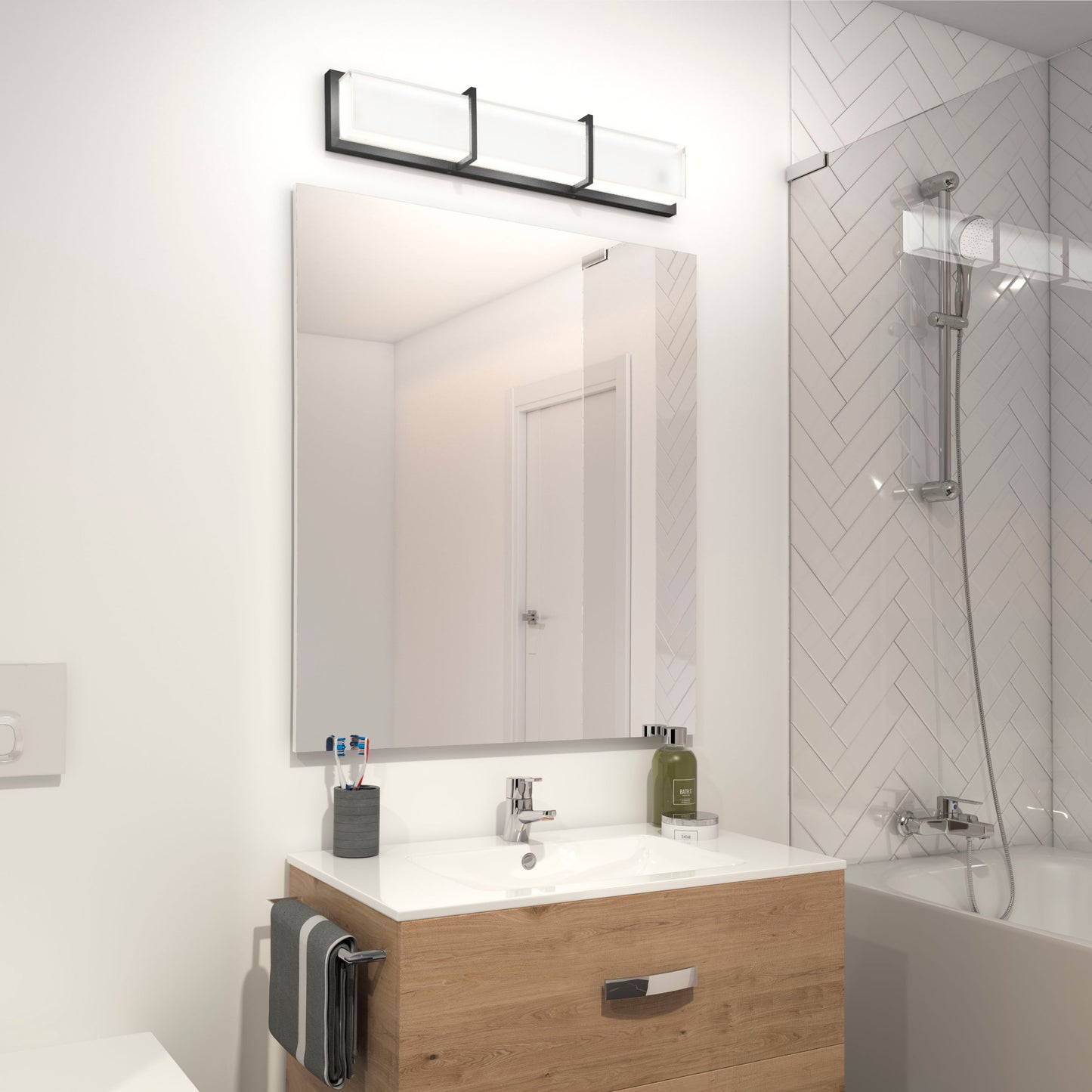 Modern bathroom featuring an Artika Subway LED vanity light above a rectangular mirror, wooden vanity, toothbrush holder, and a bathtub with glass screen against a herringbone tile wall.
