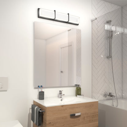 Modern bathroom featuring an Artika Subway LED vanity light above a rectangular mirror, wooden vanity, toothbrush holder, and a bathtub with glass screen against a herringbone tile wall.