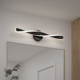 A modern LED wall light with a twisted, wave-like design in black and white is mounted above a large mirror on a white wall, with a wooden accent and a green plant visible in the corner.