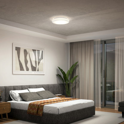 A modern, minimalist bedroom features a large bed with a gray upholstered headboard, abstract wall art, a potted plant, sheer curtains, and is illuminated by the Artika Hemisphere 15 in. LED flush mount light with 5 CCT settings.