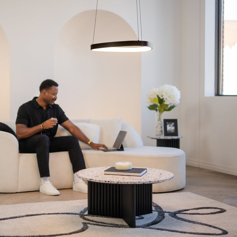 A man sits on a light-colored sofa in a modern living room, using a laptop and holding a drink. Nearby are a round coffee table and side table with white flowers. Overhead hangs the Artika Bjorn modern LED pendant light 5 CCT.